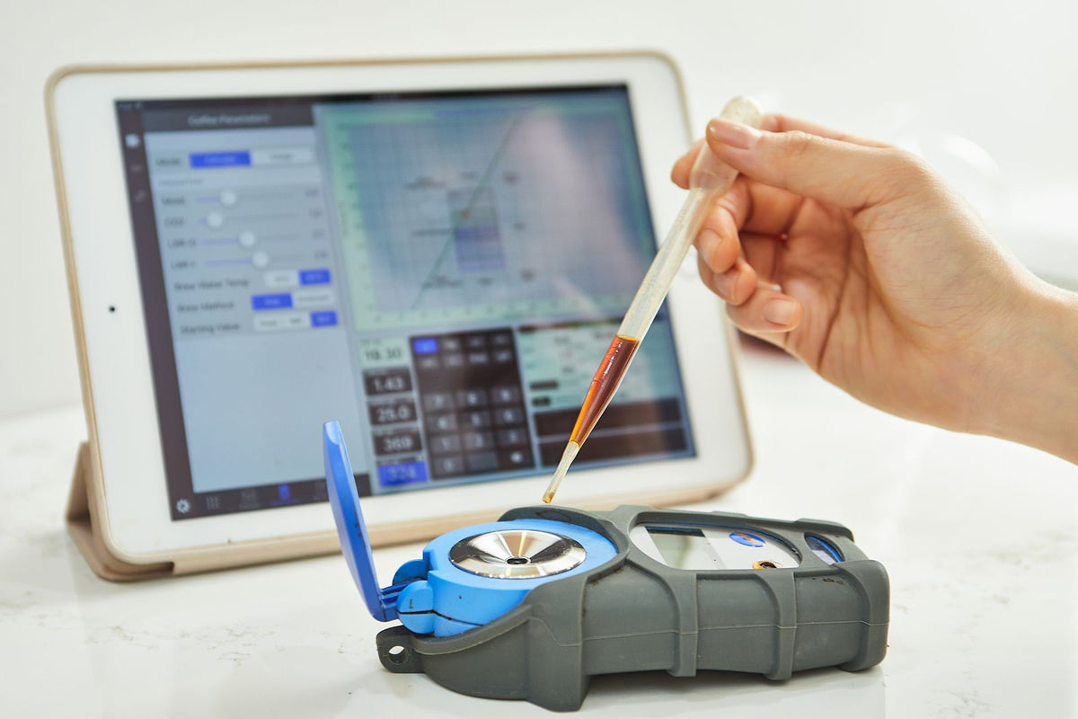 A hand holding a dropper of amber liquid over the open sample well of a handheld digital refractometer, with a tablet showing a chart behind it
