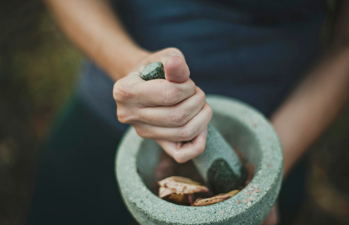 A hand grinding pieces of dried root and pink salt crystals with a pestle in a grey stone mortar.