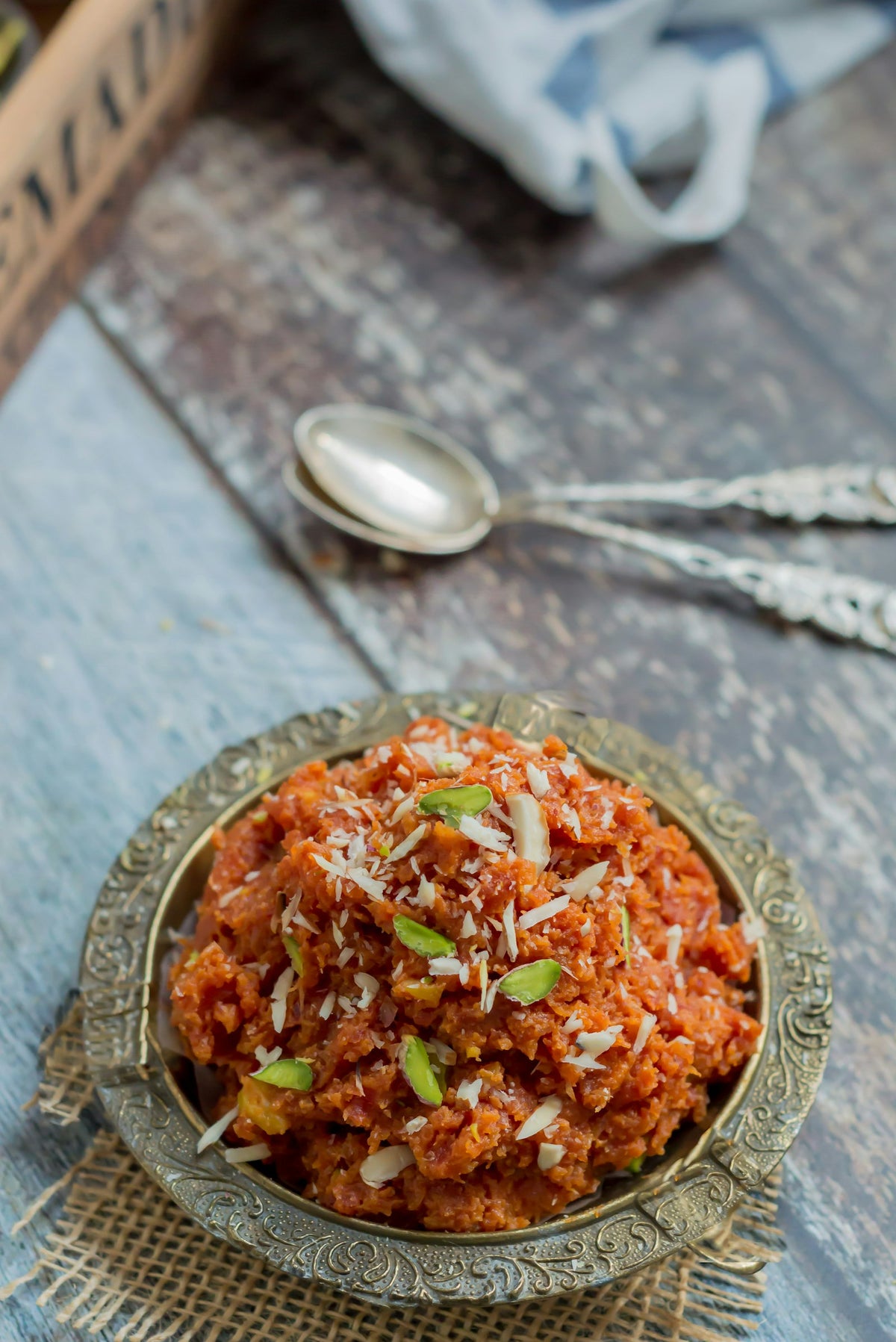 Carrot halwa topped with slivered pistachio and almond in an engraved brass bowl on a jute mat, with two silver spoons behind