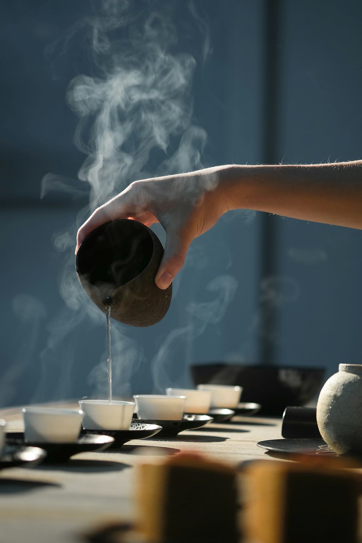 A hand pouring steaming tea from a dark pot into small white cups