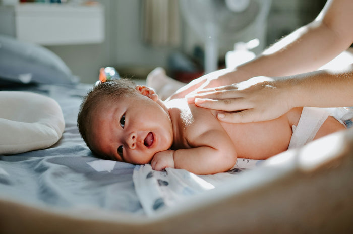 A baby in a nappy lying on its front on a grey bed sheet, an adult's hands resting on its back