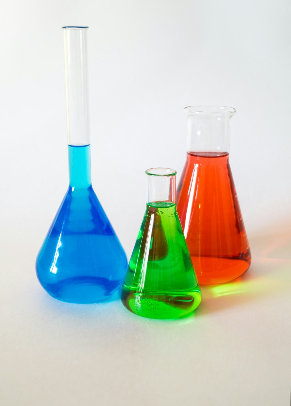 Three glass laboratory flasks holding blue, green and orange liquids on a white surface