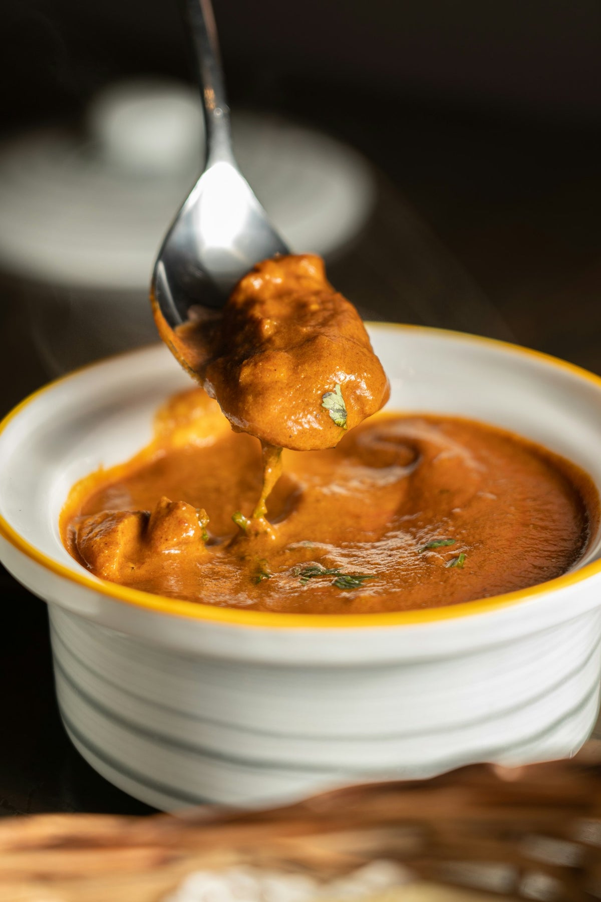 A spoon lifting a chunk coated in thick orange gravy from a white bowl with a yellow rim, with steam rising