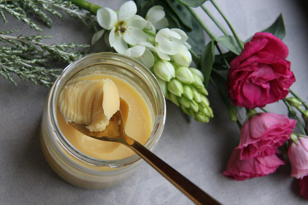 An open jar of set ghee with a spoon, beside pink flowers