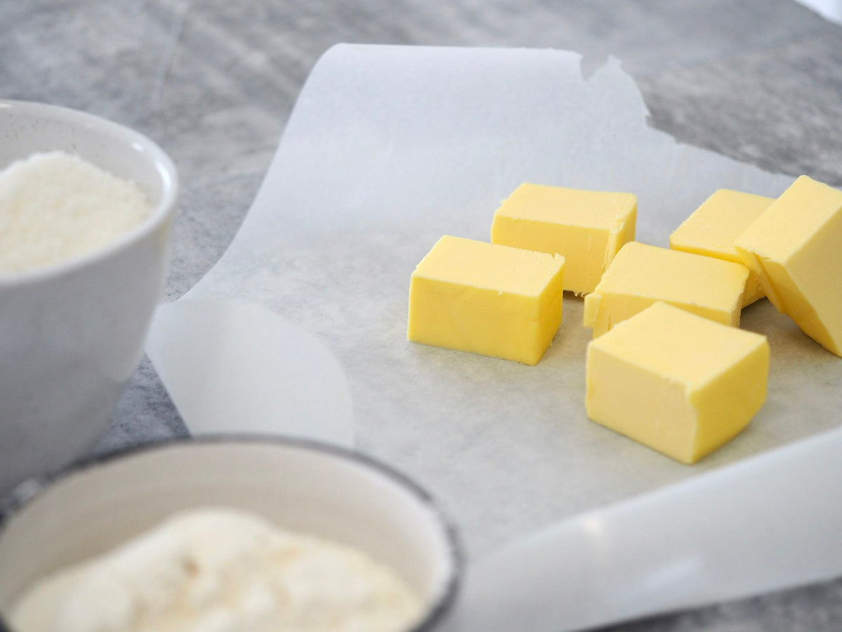 Six cubes of pale yellow butter on a sheet of baking paper, with a bowl of white sugar and a bowl of flour beside them on a grey counter