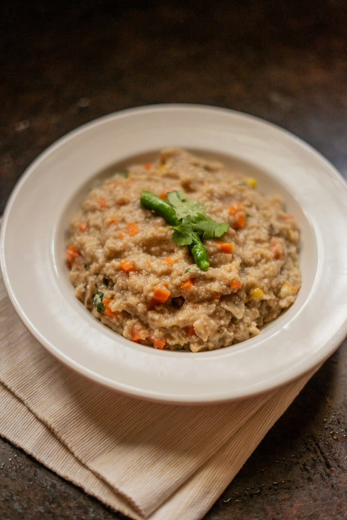A white bowl of beige semolina porridge with diced carrot, a green chilli and a coriander sprig, on a cream cloth