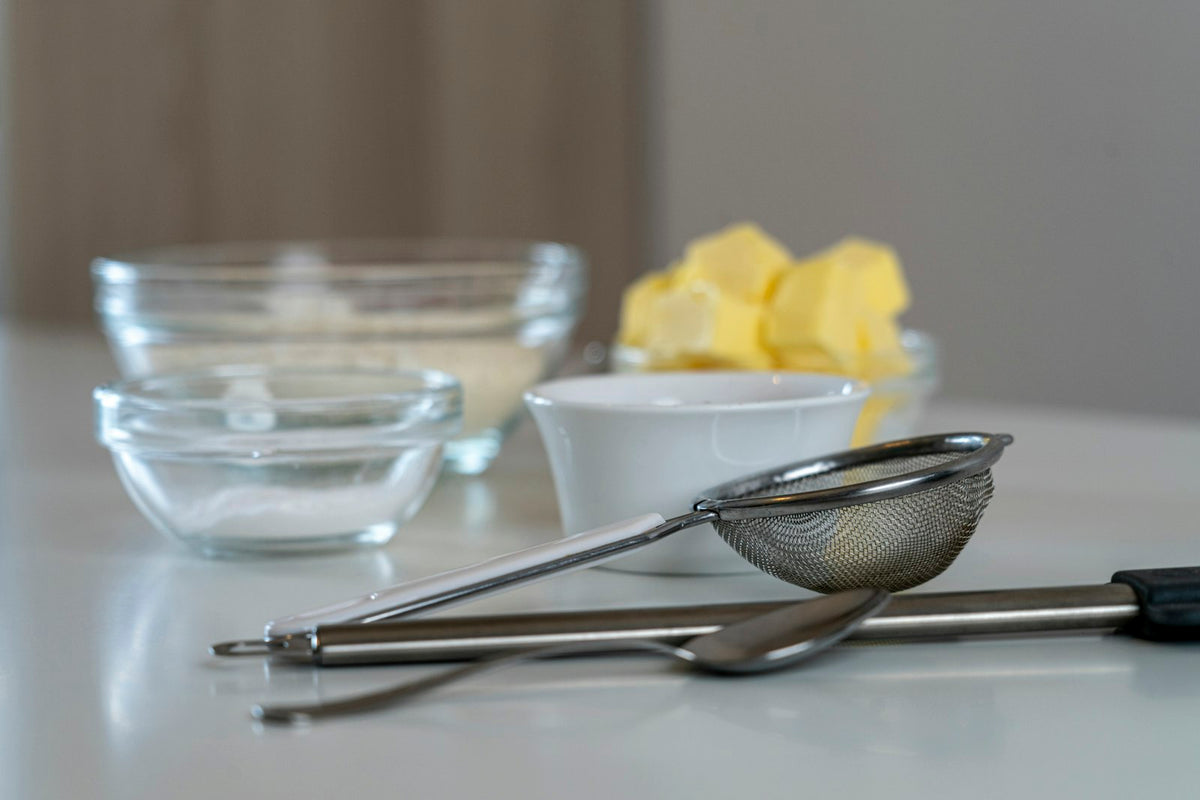 Cubes of butter in a white bowl behind a fine mesh strainer, with glass bowls on a white counter