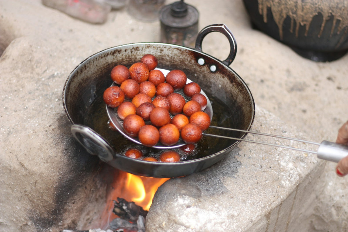 Round golden-brown gulab jamun being lifted from hot oil on a slotted ladle over a black iron kadai set on a wood fire