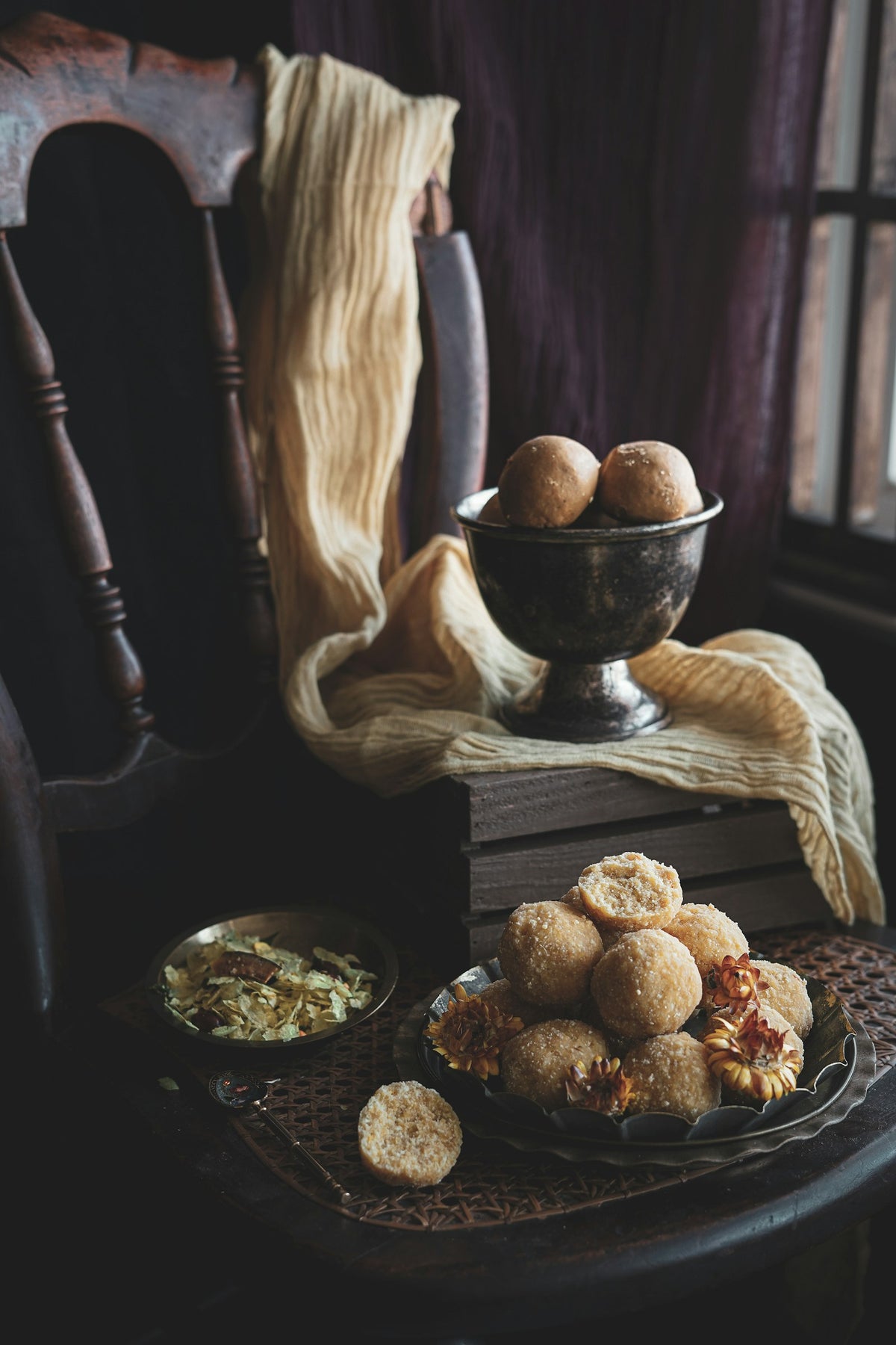 Pale round ladoos piled on a scalloped plate and in a silver footed bowl on a cane chair, with a small plate of poha beside them