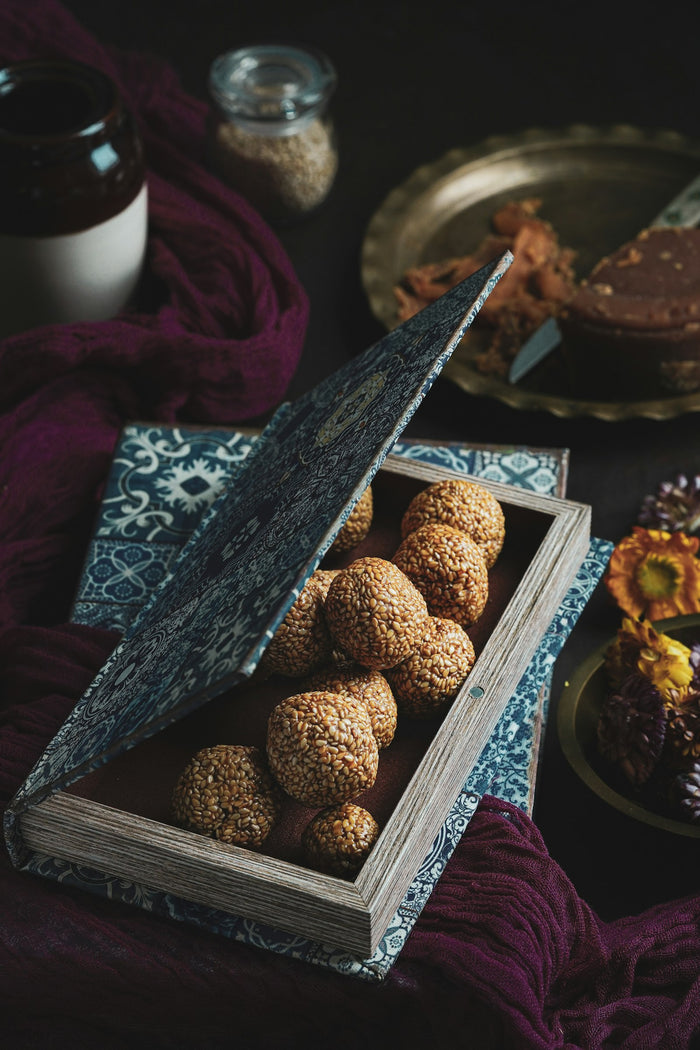 Round sesame ladoos in an open patterned box, with a jar of seeds and a plate of jaggery behind