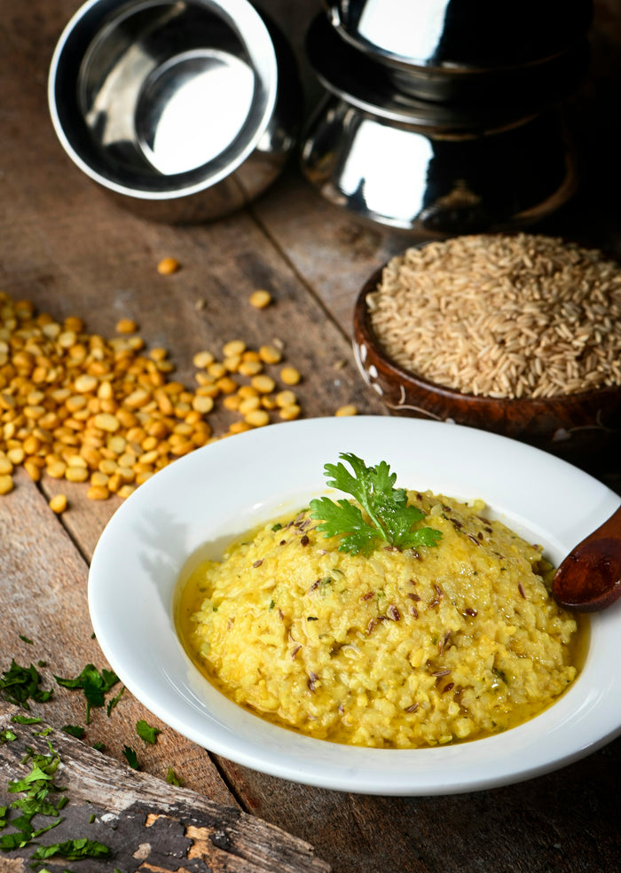 A white plate of yellow rice and lentils topped with a coriander sprig, with split dal, raw rice and steel vessels behind