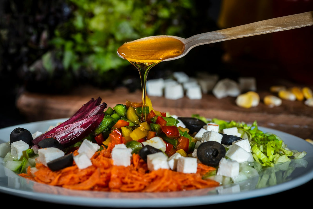 Golden oil pouring from a wooden spoon onto a plate of chopped peppers, grated carrot, beetroot, black olives, cubes of white cheese and shredded lettuce
