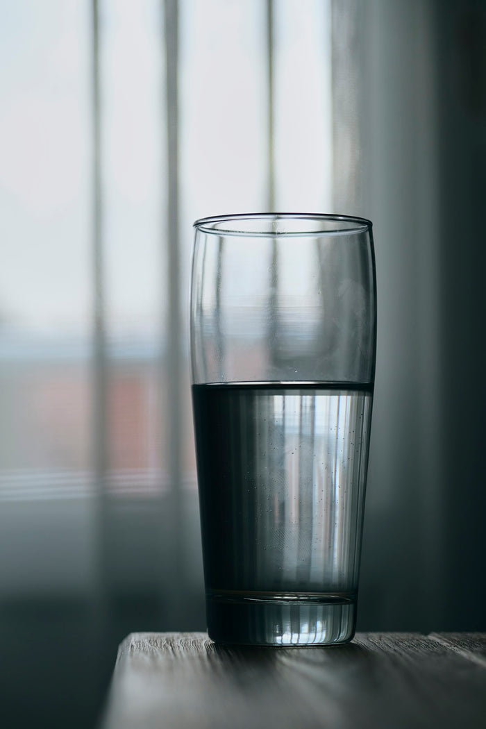 A glass half full of water on a wooden table in front of a window with pale curtains