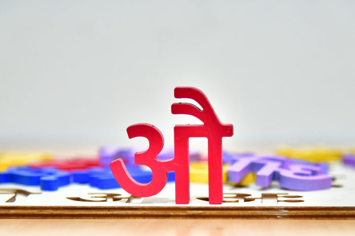 A red wooden Devanagari letter standing upright on a wooden puzzle board, with blurred blue, yellow and purple letters behind