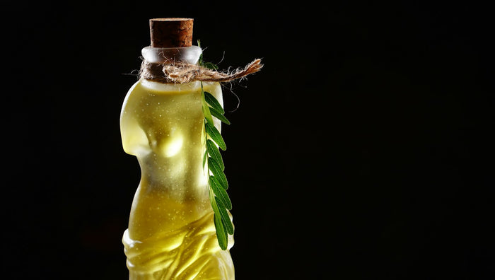 A sculpted glass bottle of golden oil with a cork stopper and a sprig of leaves, on a black background