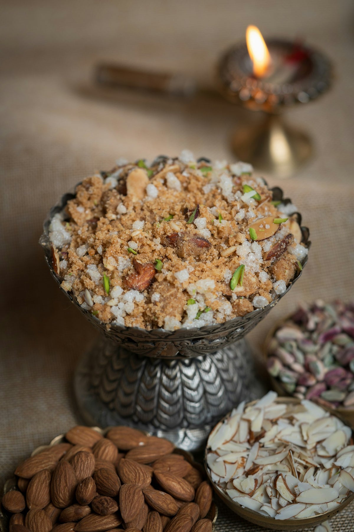 A crumbly brown wheat sweet with sugar crystals, almond slivers and pistachio in a silver footed bowl, with almonds and a lit diya beside it