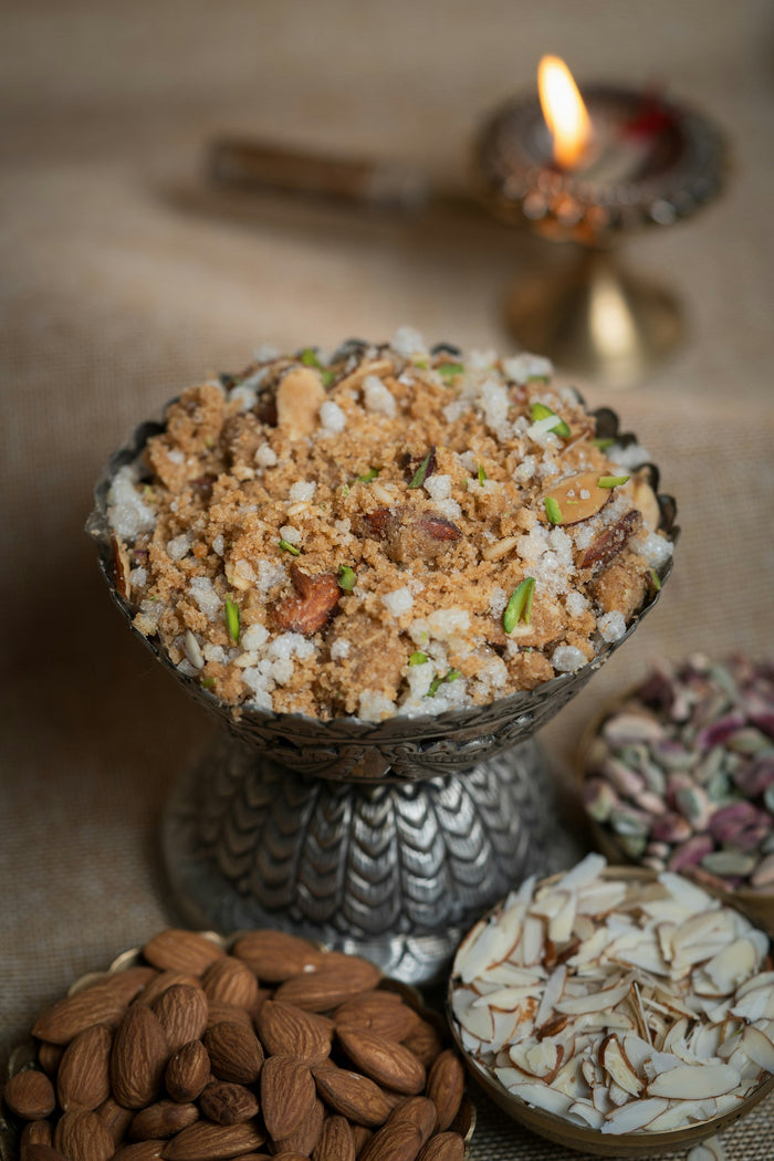 A crumbly brown wheat sweet with sugar crystals, almond slivers and pistachio in a silver footed bowl, with almonds and a lit diya beside it