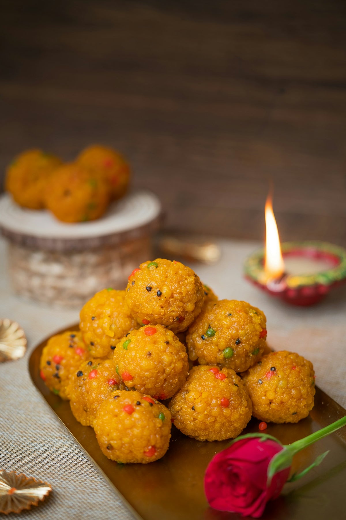 A pile of orange boondi ladoos flecked with red and green on a brass tray, with a lit clay diya and a red rose behind