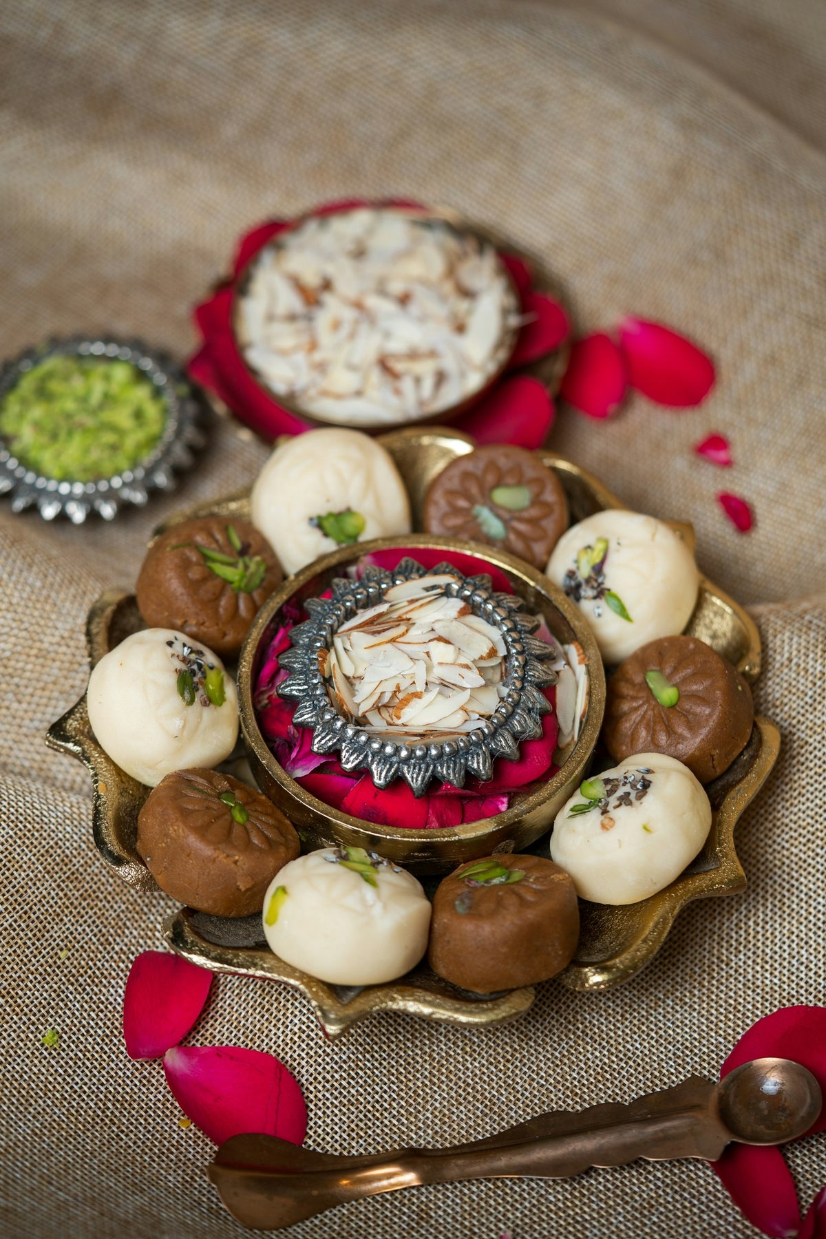 White and brown milk sweets topped with pistachio on a brass tray, around a bowl of sliced almonds and rose petals