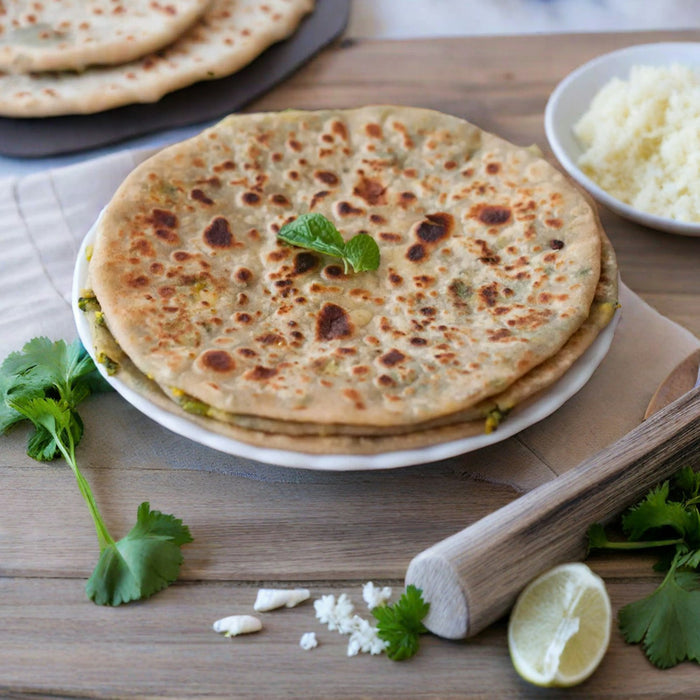 Two stuffed parathas stacked on a white plate with a mint sprig, coriander leaves, a rolling pin and half a lime on a wooden table
