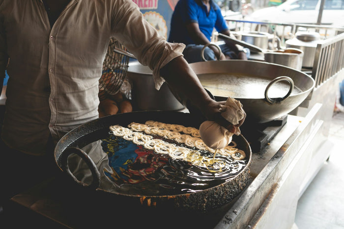 A street vendor squeezing jalebi batter from a cloth into a wide iron pan of hot oil, with rows of pale spirals frying and a second pan of syrup behind