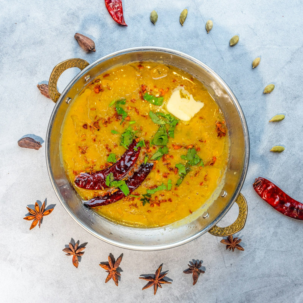 A steel kadai of yellow dal topped with fried dried red chillies, chopped coriander and a pat of butter, ringed by cardamom pods, star anise and red chillies