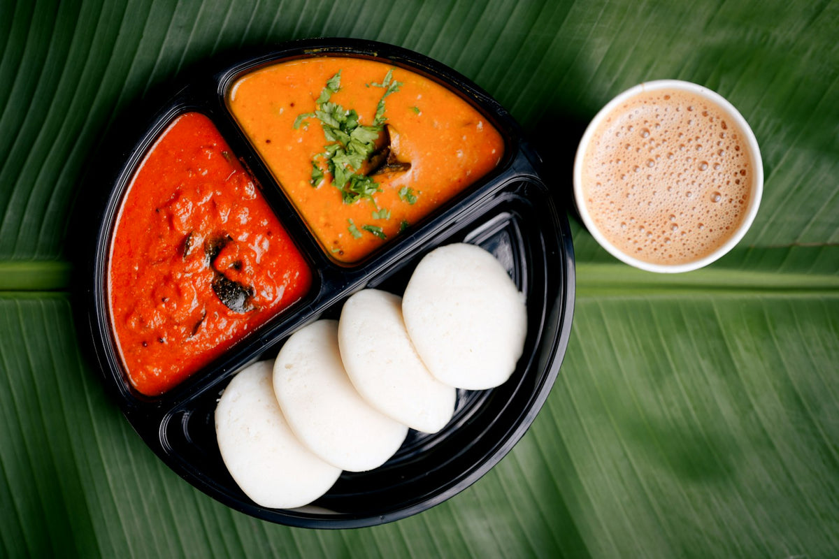 Four white idlis in a black tray with red chutney and sambar, and a paper cup of filter coffee, on a banana leaf