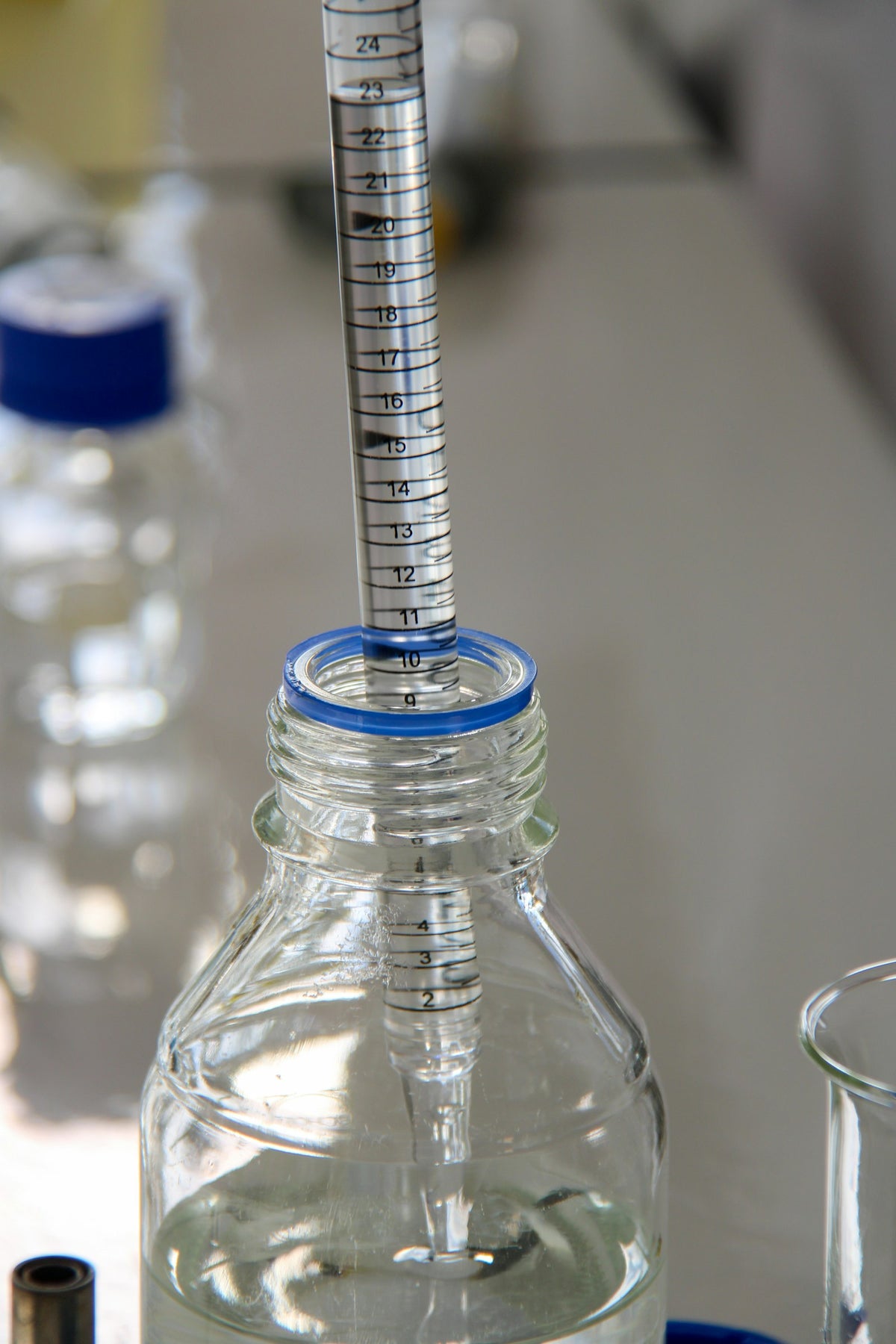 A graduated glass pipette standing in a clear reagent bottle with a blue rim on a laboratory bench, a beaker beside it