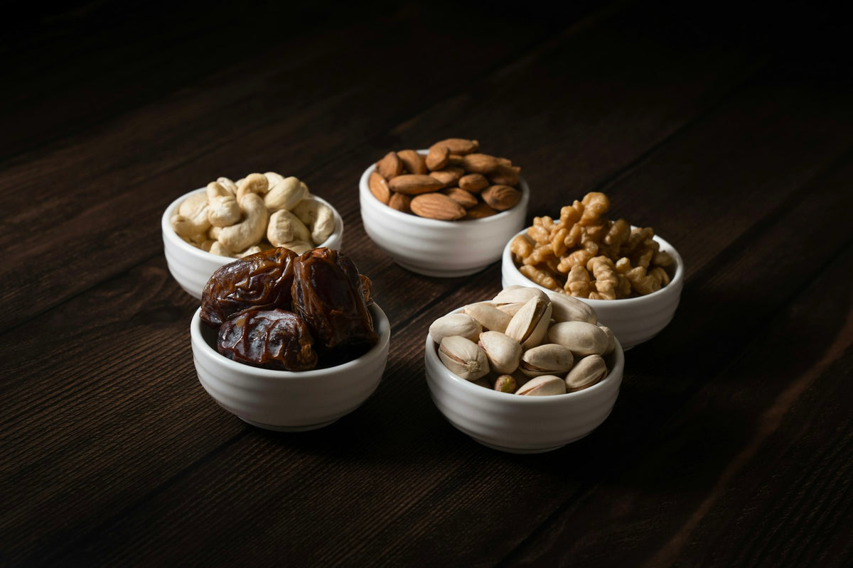 Five small white bowls of cashews, almonds, walnuts, pistachios and dates on a dark wooden table