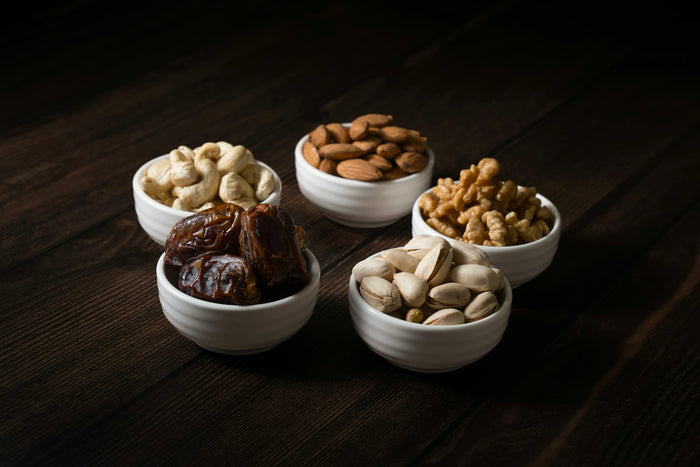 Five small white bowls of cashews, almonds, walnuts, pistachios and dates on a dark wooden table