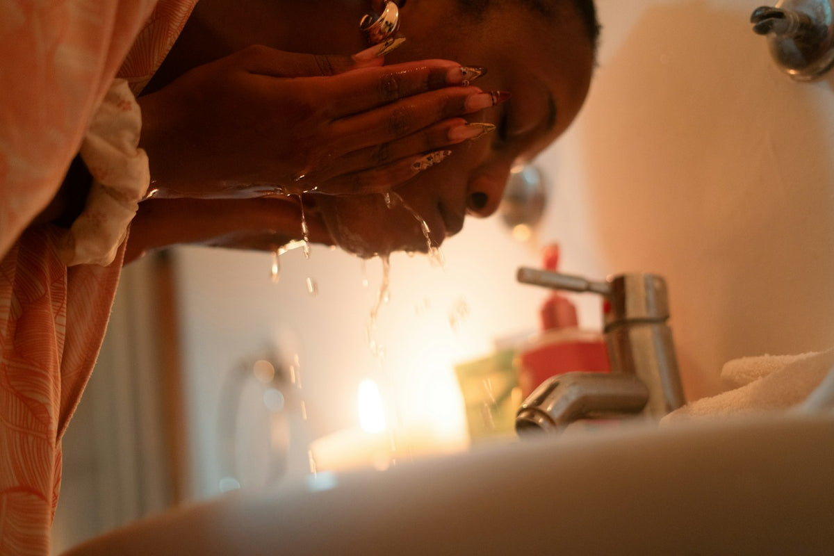 A woman bent over a bathroom sink splashing water onto her face with both hands, a candle glowing behind the tap