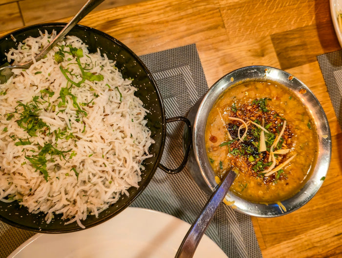 A black bowl of jeera rice scattered with coriander beside a steel bowl of tempered yellow dal, each with a spoon, on a wooden table